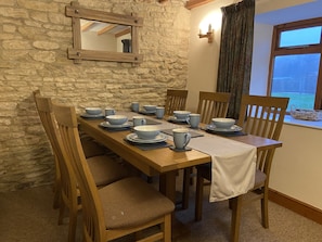 Une table à manger dressée avec des bols et des assiettes bleus, des chaises en bois, un mur en pierre, une fenêtre avec des rideaux et une poutre en bois au plafond.