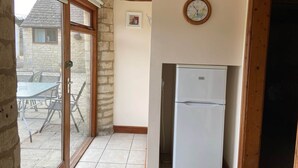 A kitchen with a white refrigerator, a wooden door frame, and a window with a view of a stone building and outdoor furniture. - Stable Cottage, Old Mill Farm, Cotswold Water Park (Cirencester)