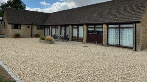 A stone building with a gravel courtyard, featuring a dining area with chairs and tables, and a large window with curtains. - Stable Cottage, Old Mill Farm, Cotswold Water Park (Cirencester)