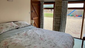 A bedroom with a large bed, floral bedding, a wooden door, and a view of a gravel area with outdoor furniture. - Stable Cottage, Old Mill Farm, Cotswold Water Park (Cirencester)