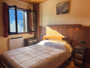 Une chambre à coucher avec un lit, une fenêtre avec des rideaux, un radiateur, une tête de lit en bois, une table de chevet et un tableau accroché au mur.