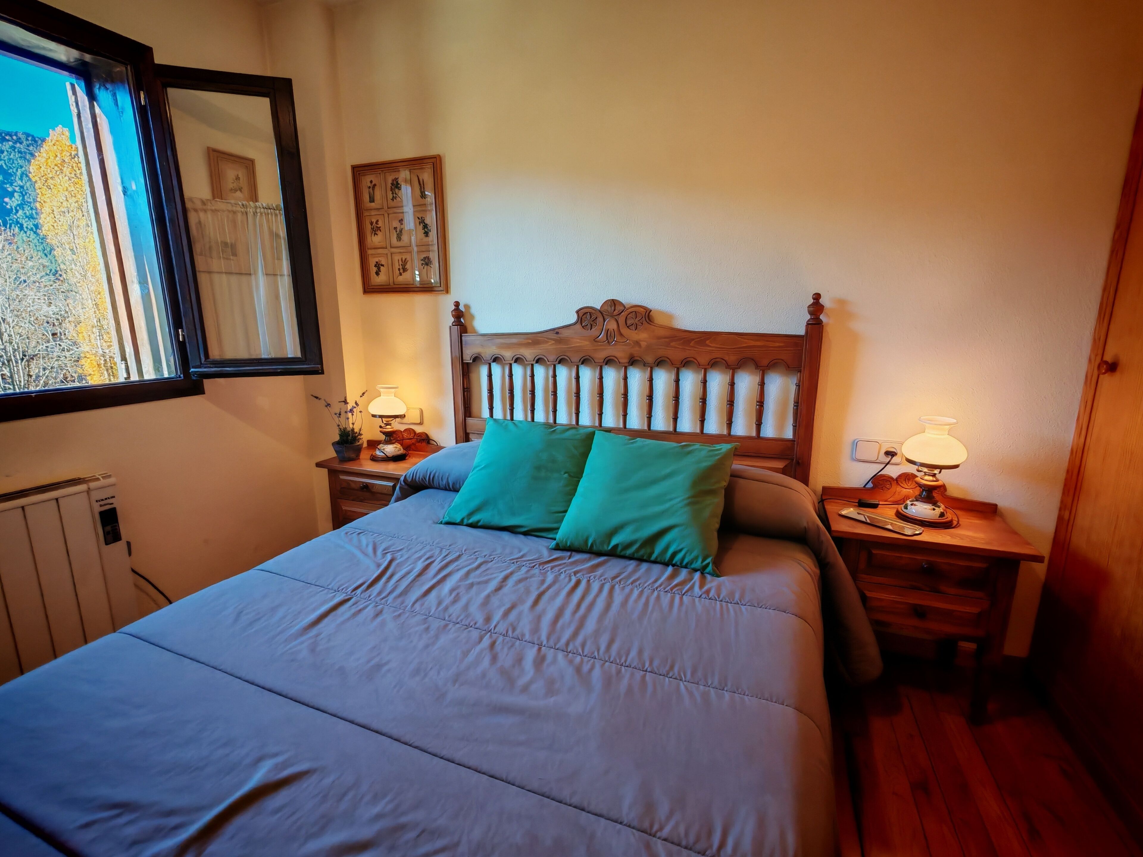 Une chambre à coucher avec une tête de lit en bois, un lit recouvert d’une couverture bleue, deux tables de chevet avec des lampes, une fenêtre donnant sur des arbres et un tableau accroché au mur.