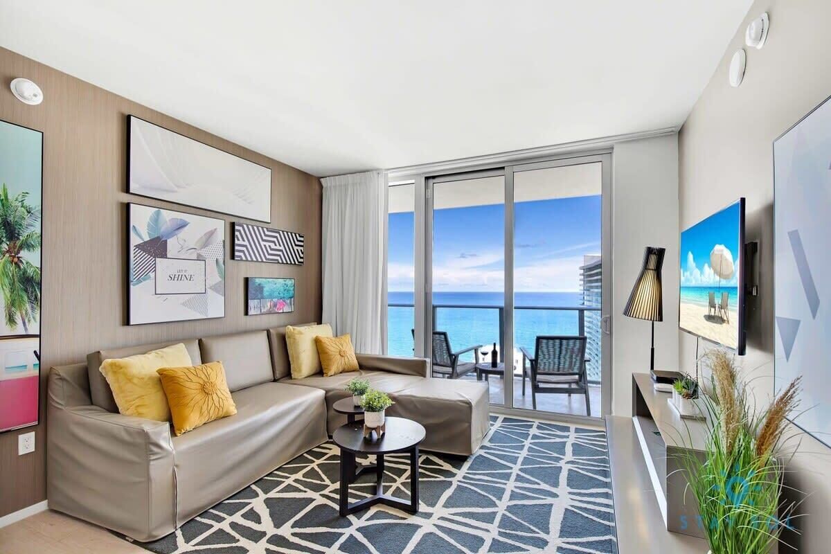 A modern hotel room with a large balcony, a sofa, a coffee table, and artwork on the walls.
