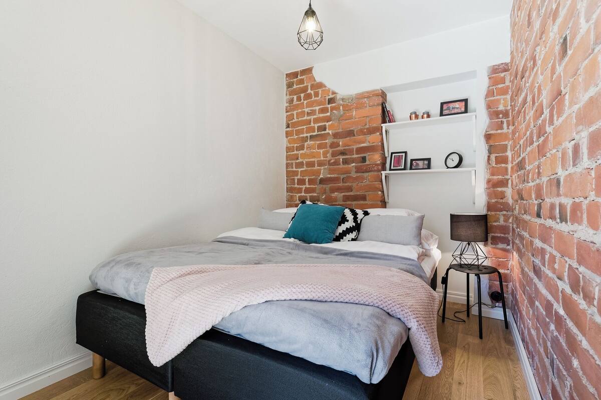 A bedroom with a brick wall, a bed with a grey and black headboard, a white shelf with framed photos, and a small table with a lamp.