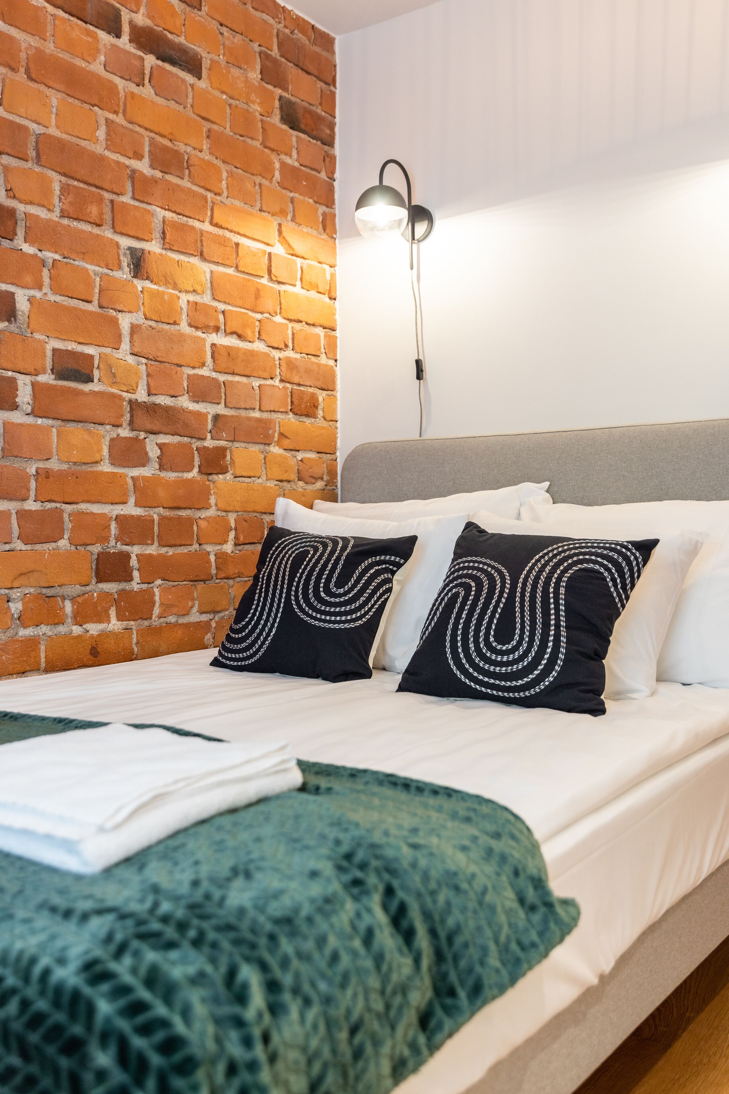 A hotel room with a brick wall, a bed with white bedding, two black pillows with white patterns, and a bedside lamp.