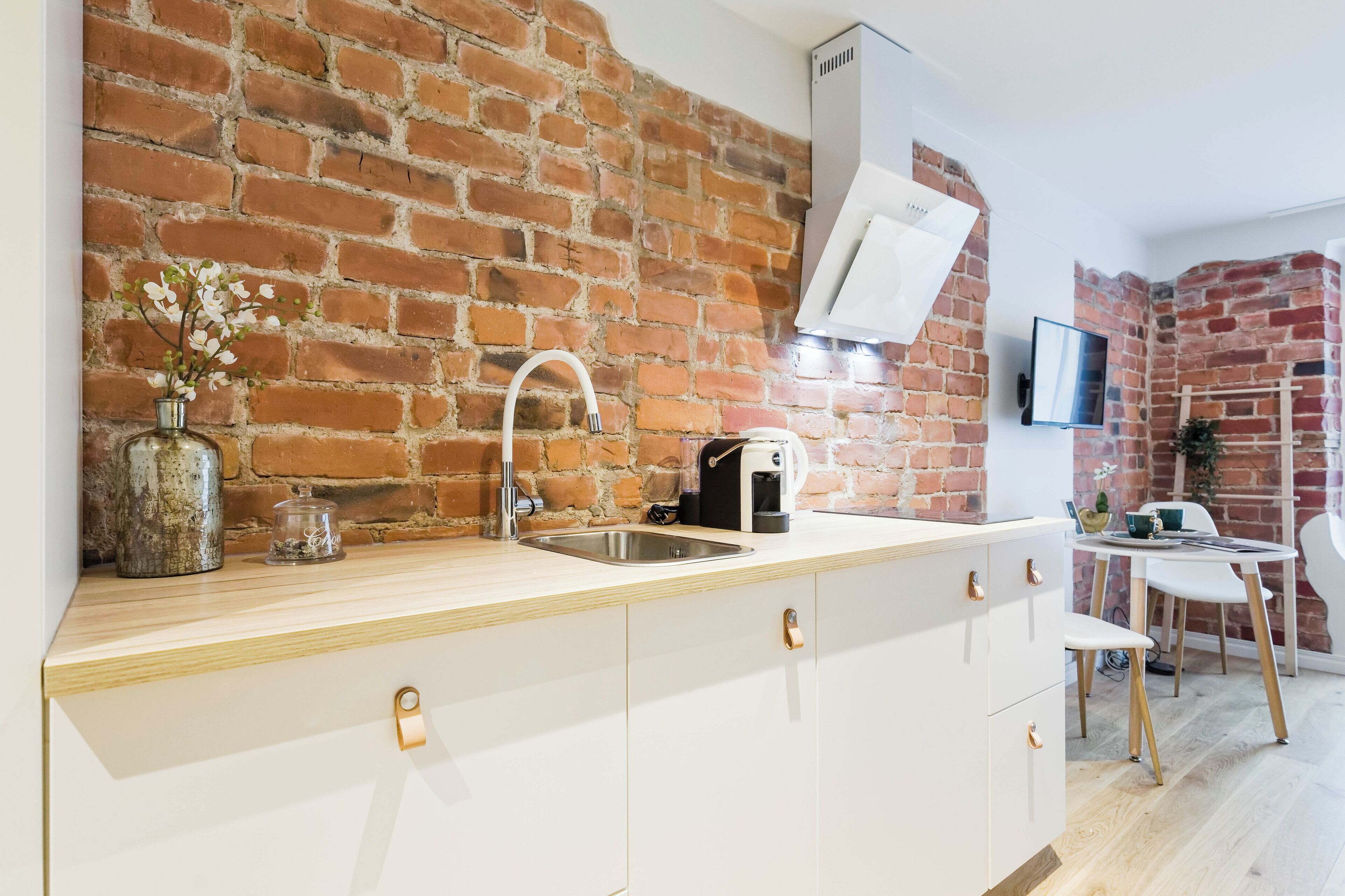 A kitchen with white cabinetry, a brick wall, a stainless steel sink, a kettle, and a small table with chairs.