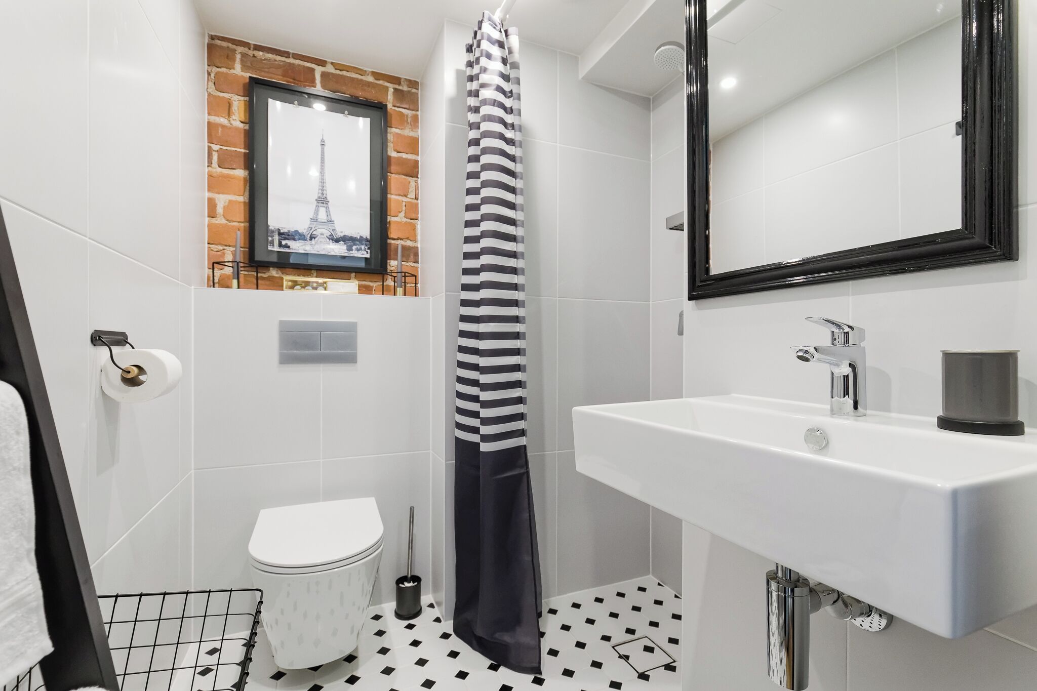 A bathroom with a white sink, a toilet, a shower with a striped curtain, a mirror, and a picture of the Eiffel Tower on the wall.