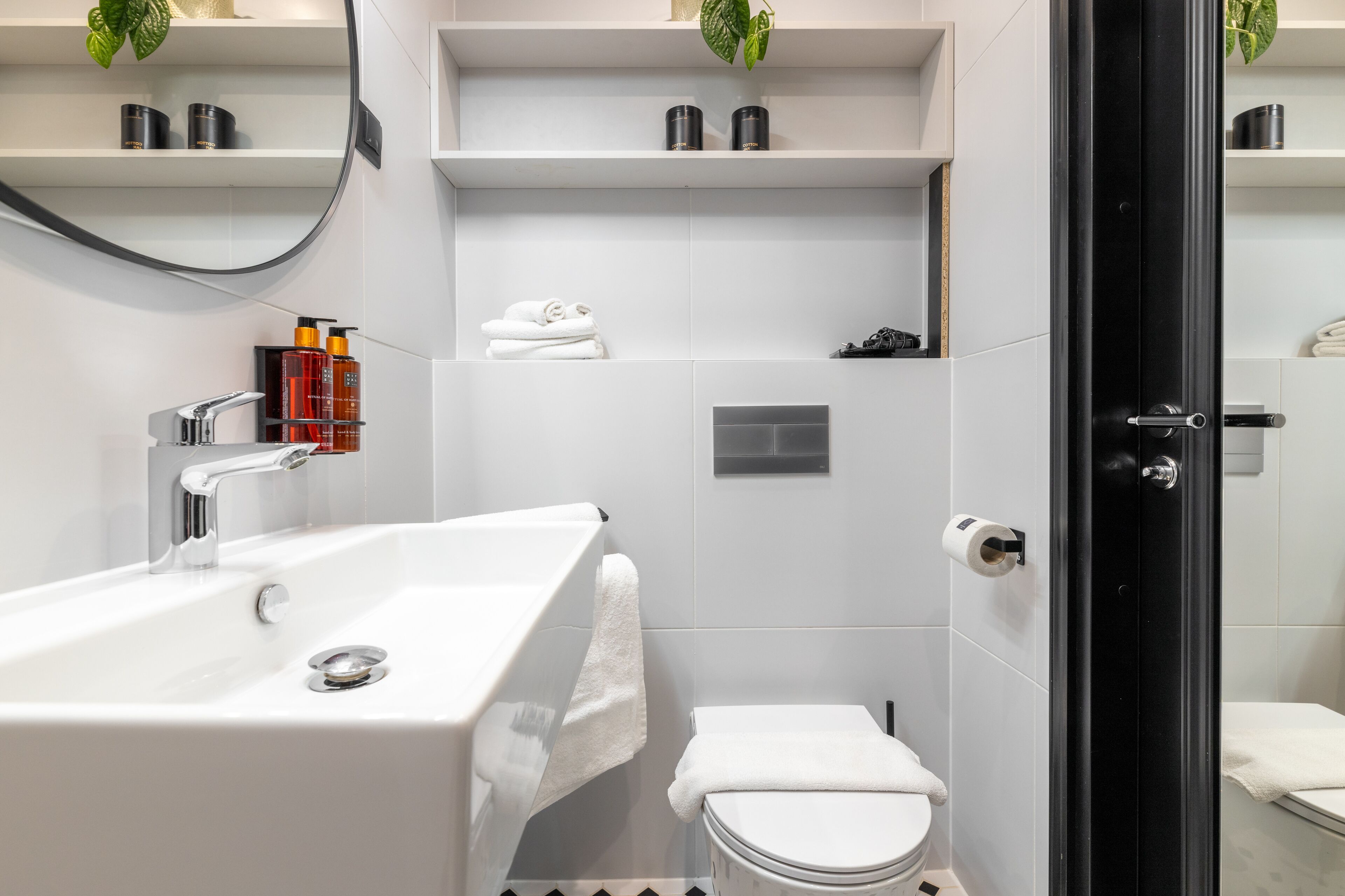 Un bagno moderno con un ampio lavandino bianco, un water e una zona doccia. Si notano mensole con articoli da toilette e una pianta.