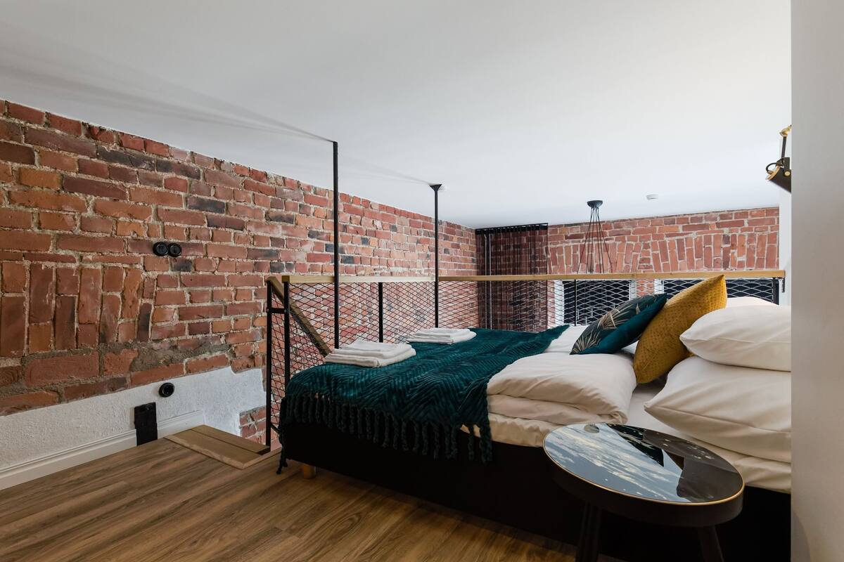 A room with a brick wall, a bed with white and green bedding, a small table, and a hanging light fixture.