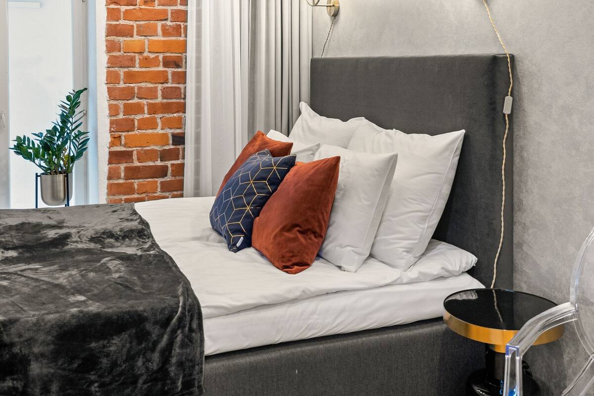 A modern bedroom with a brick wall, a grey bed, white bedding, and a small side table.