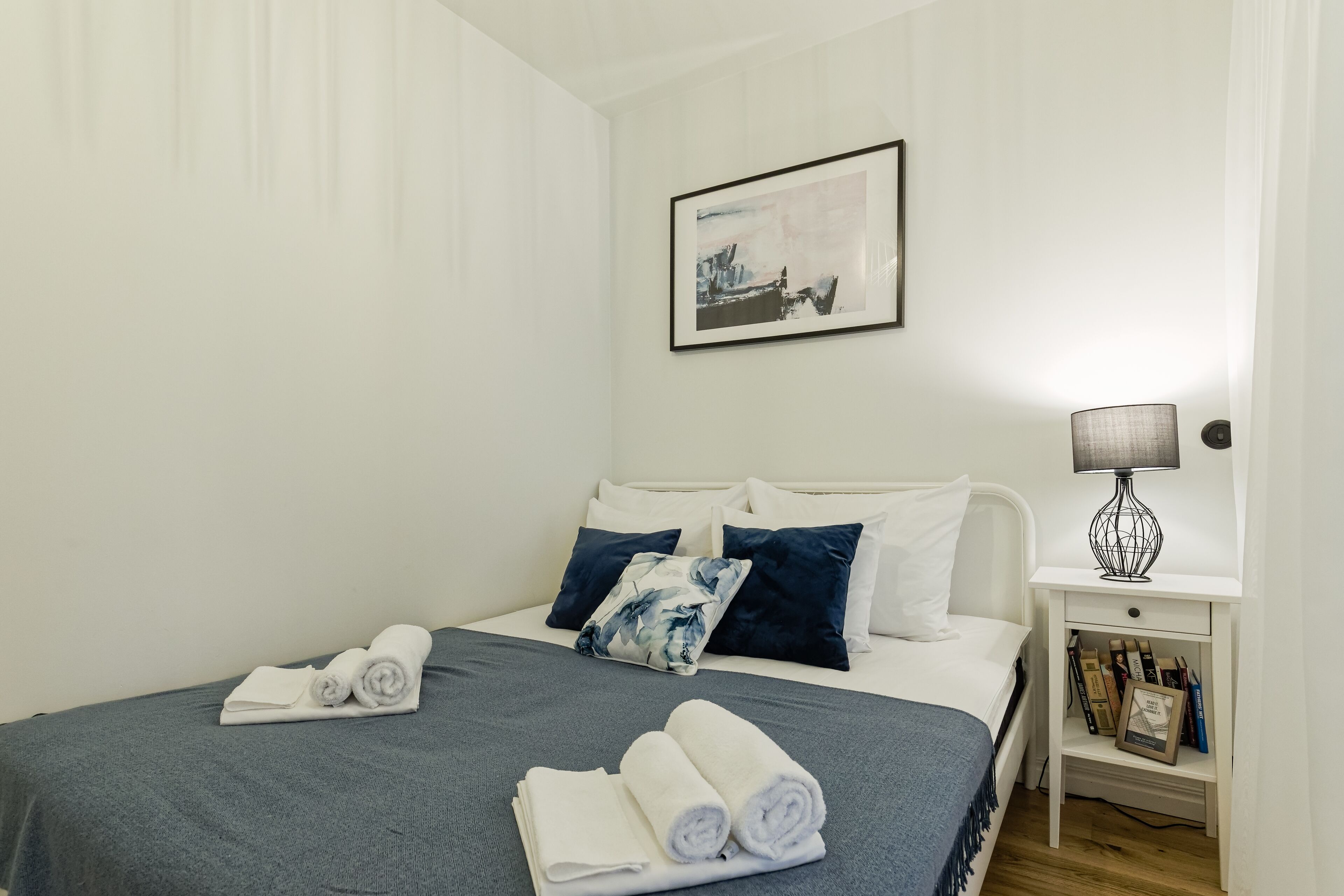 Una cama bien hecha con toallas enrolladas, una mesita de noche con una lámpara y un cuadro en la pared.