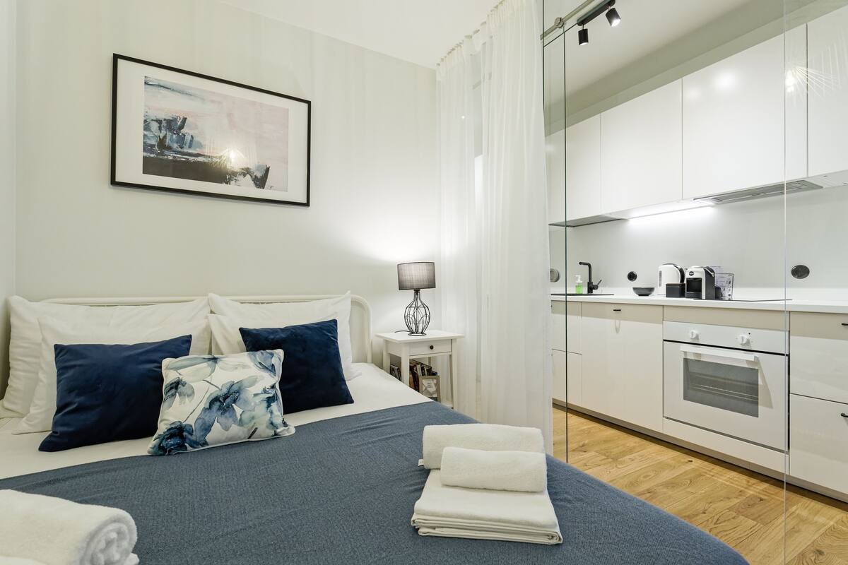 A modern hotel room with a neatly made bed, a small bedside table with a lamp, a kitchen area with a microwave and toaster, and a framed picture on the wall.