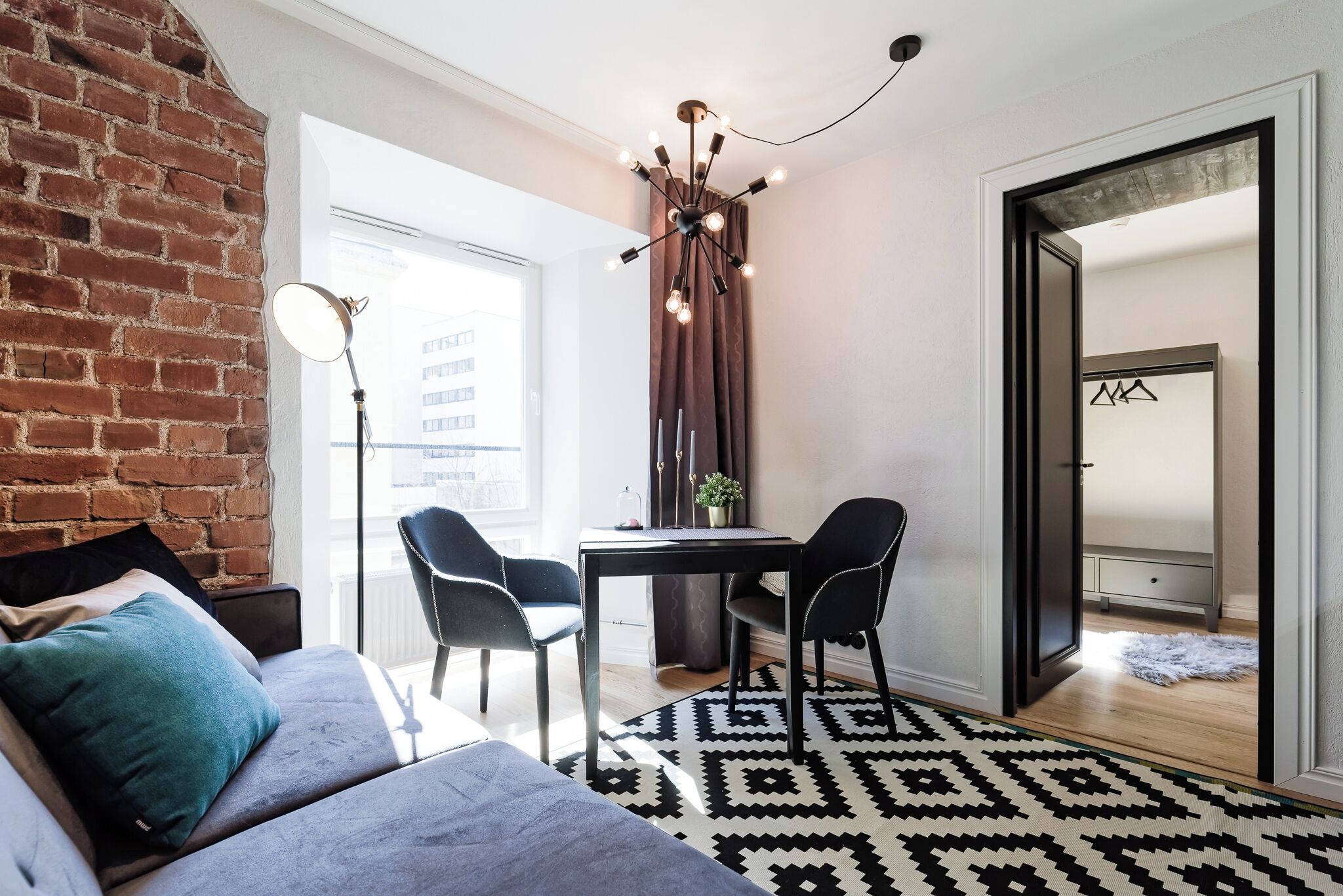 A modern living room with a brick wall, a sofa, a dining table with chairs, a chandelier, and a view through an open door.
