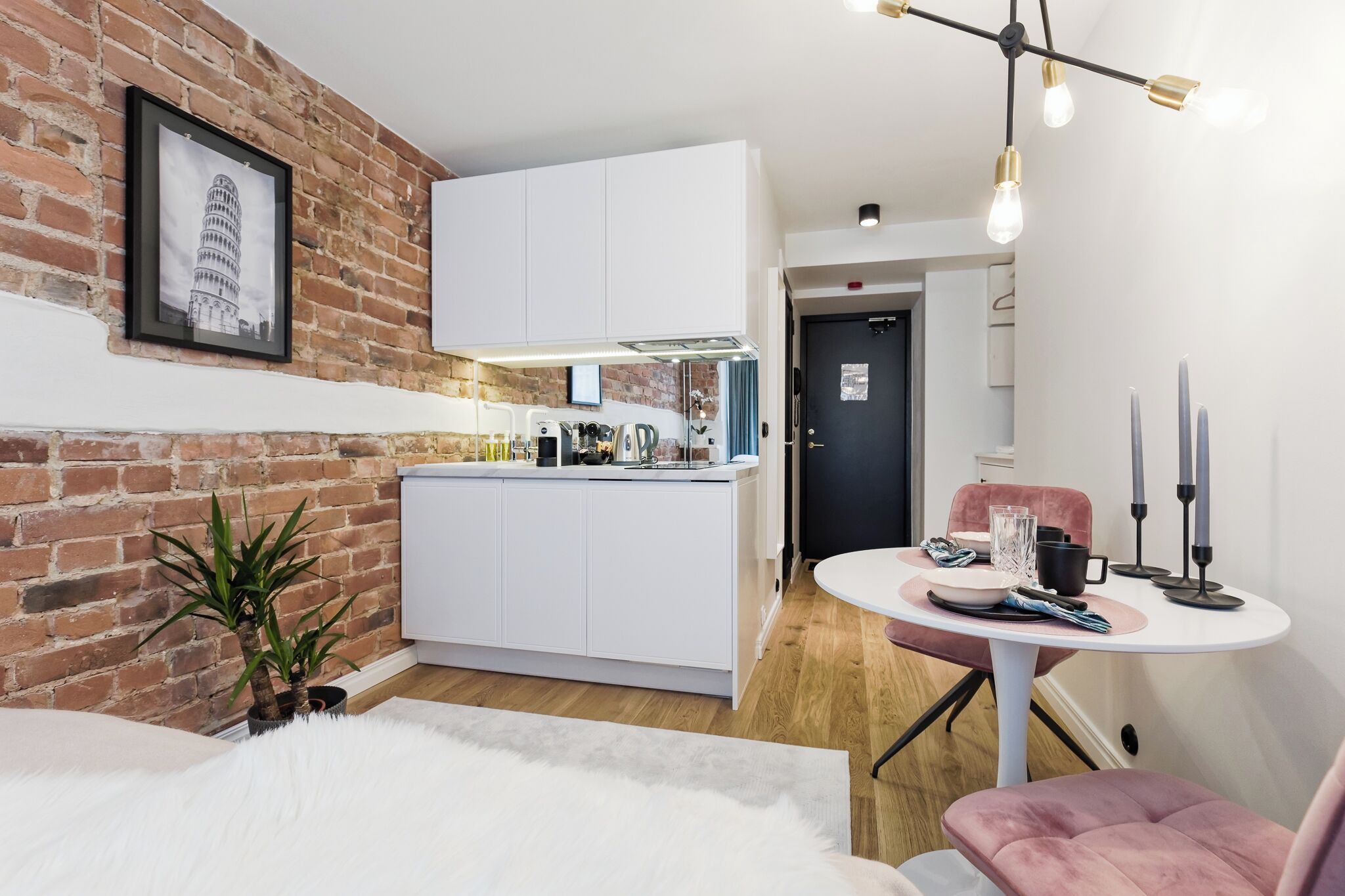 Cocina compacta con armarios blancos, pared de ladrillo y zona de comedor con mesa redonda y silla rosa.