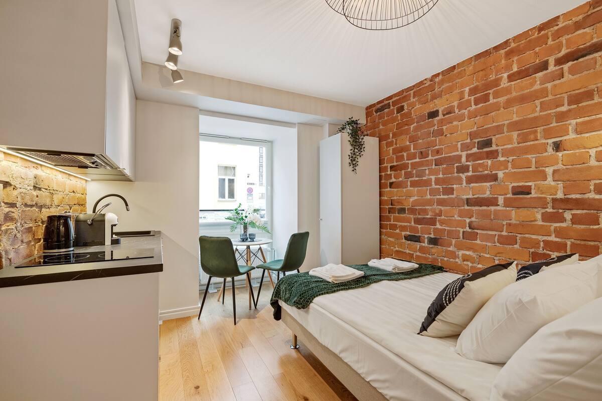 A modern room with a brick wall, a bed with white bedding, a small kitchenette, and a view through a window.