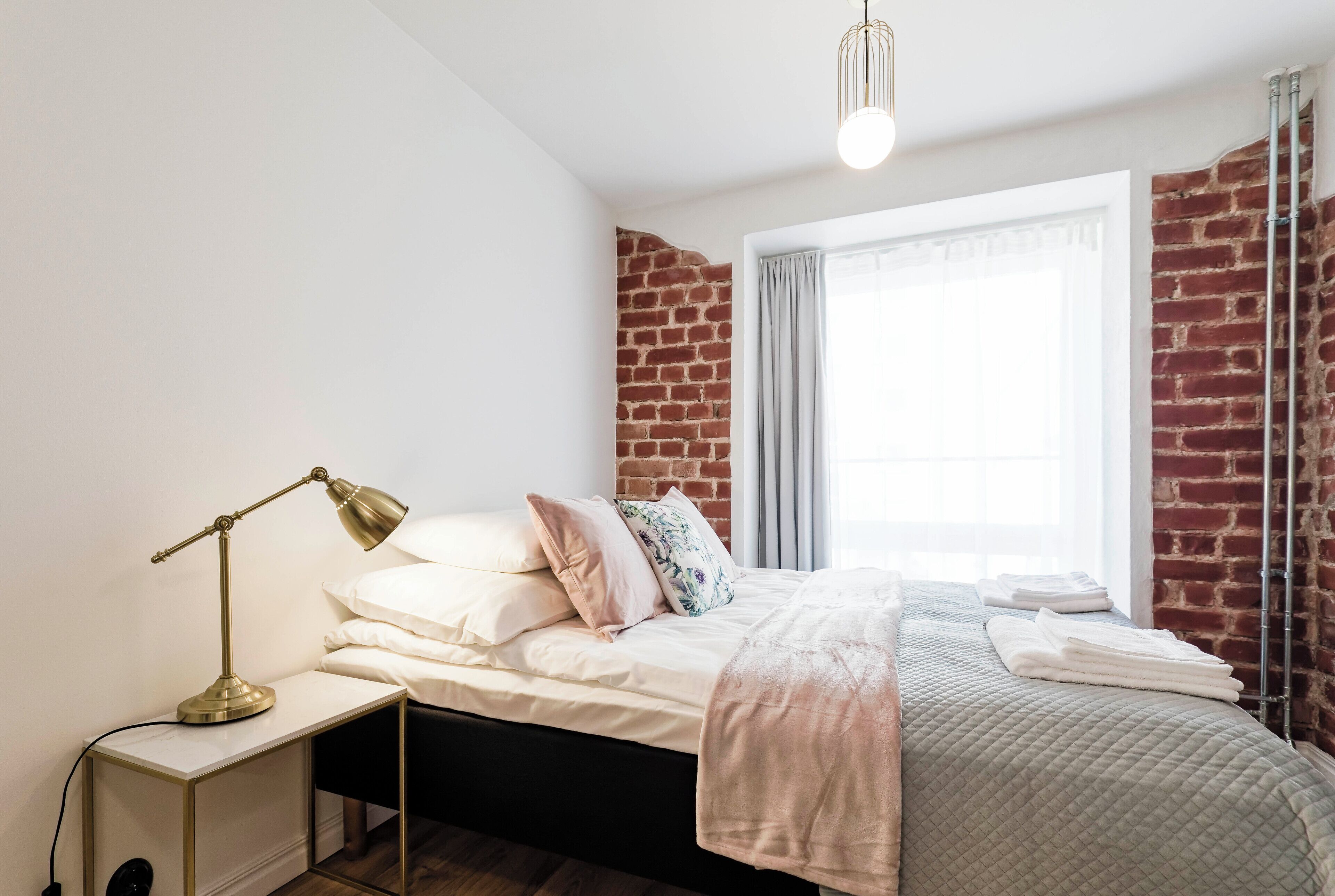 Ein Schlafzimmer mit Backsteinwand, einem Bett mit weißer Bettwäsche, einer Nachttischlampe und einem Fenster mit Vorhängen.