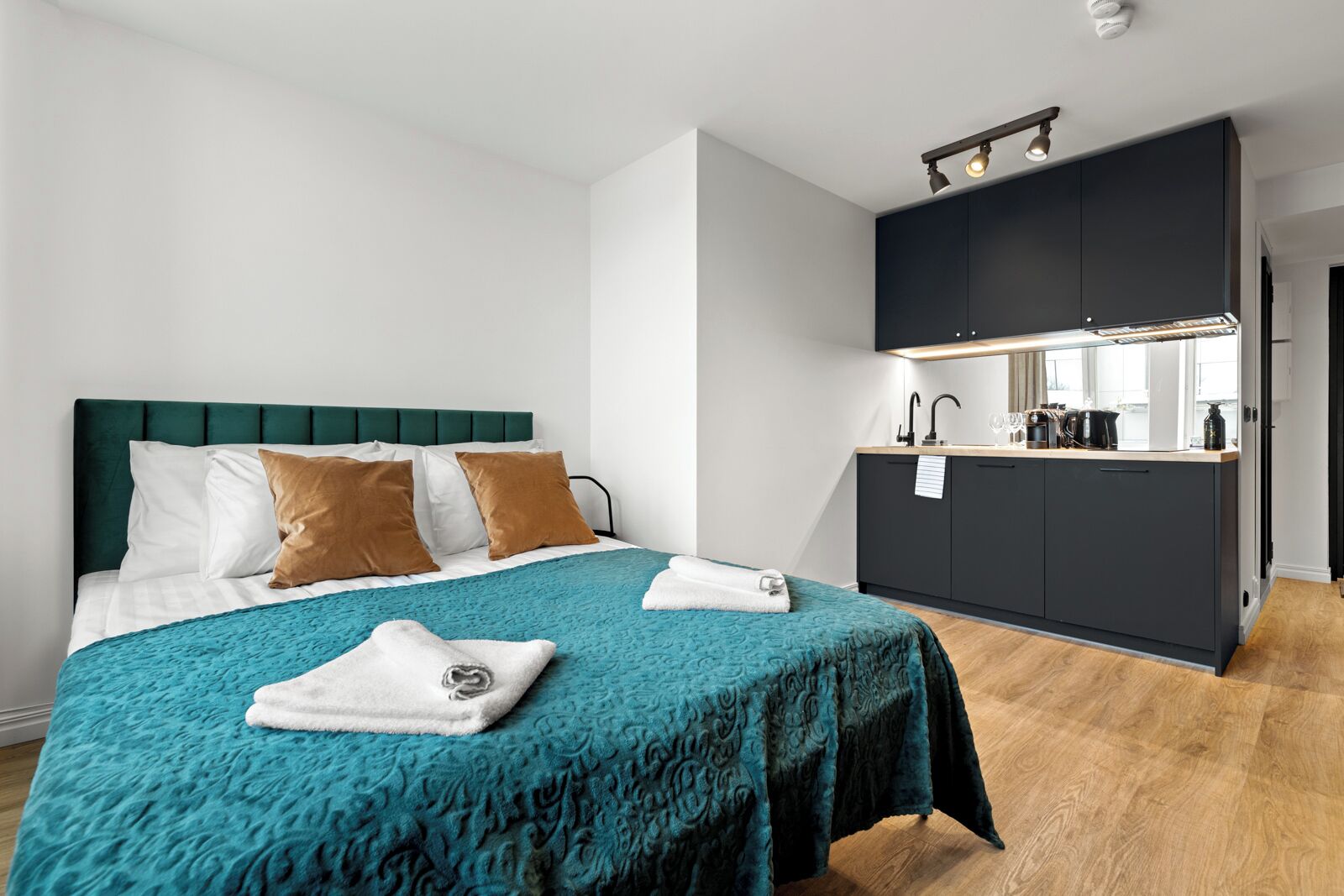 Une chambre d’hôtel moderne, dotée d’un lit, d’une couvre-lit de couleur turquoise et d’une kitchenette avec des meubles de couleur sombre et un plan de travail.