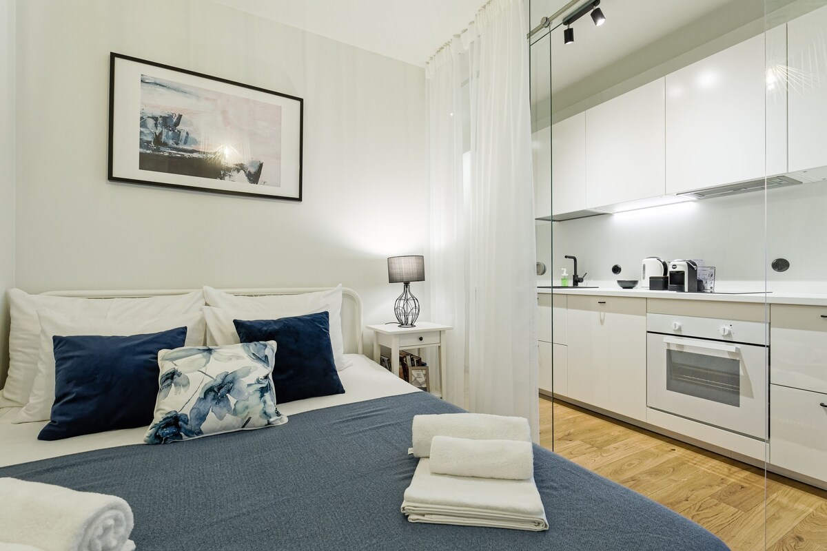 A modern hotel room with a bed, a bedside table with a lamp, a kitchen area with a stove, and a framed picture on the wall.