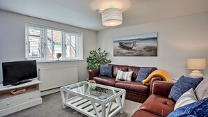A living room with a brown leather sofa, a glass coffee table, a television on a white stand, a painting of a boat on the wall, and a ceiling light fixture. - Old Salt Loft (Whitby)