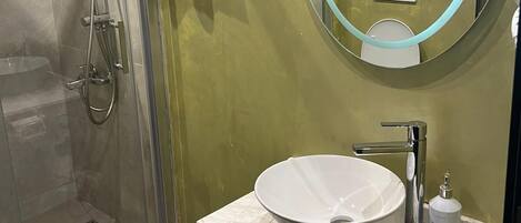 A modern bathroom with a glass-enclosed shower, a white sink with a round mirror, and a green wall.
