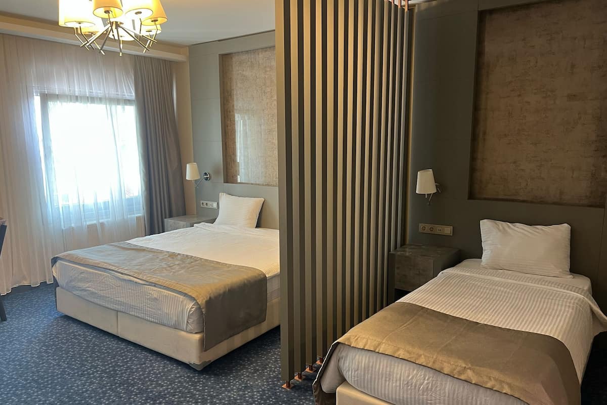 Quarto de hotel com duas camas, uma janela grande com cortinas, um lustre e uma parede com textura.