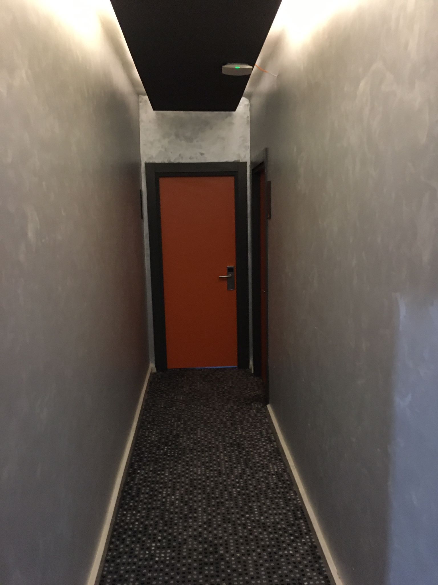 Um corredor estreito com piso coberto por carpete, uma porta vermelha no final e paredes em tom claro.