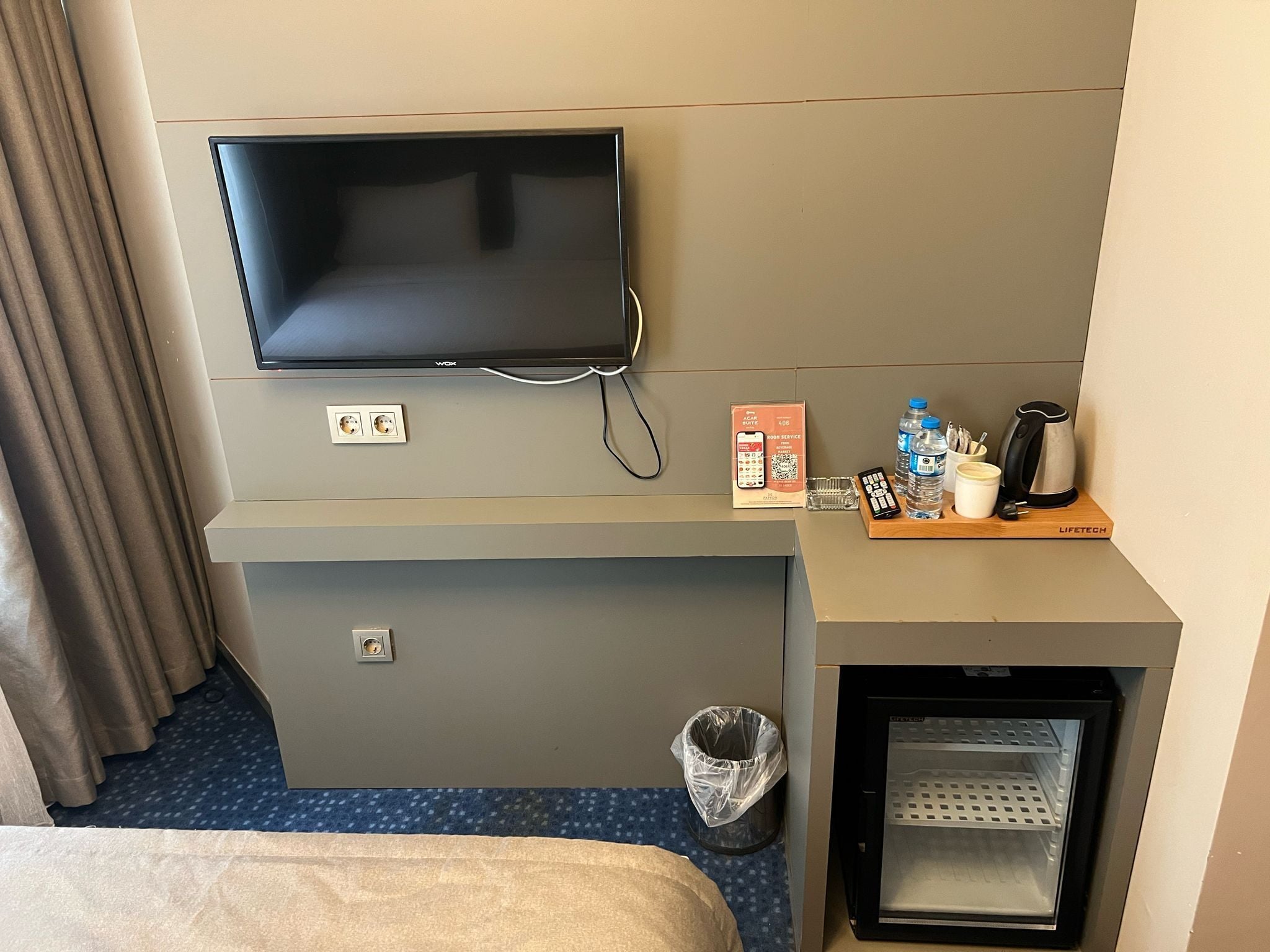 Habitación de hotel con televisión de pantalla plana montada en la pared, un refrigerador pequeño, un cubo de basura, una cafetera y un control remoto sobre una bandeja de madera.
