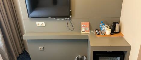 Habitación de hotel con televisión de pantalla plana montada en la pared, un refrigerador pequeño, un cubo de basura, una cafetera y un control remoto sobre una bandeja de madera.
