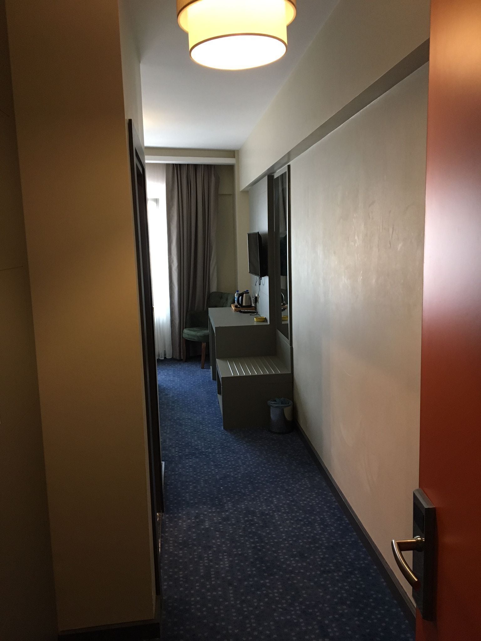 A hotel hallway with a blue carpet, a desk with a chair, a television, and a door on the right.