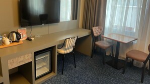 A hotel room with a flat-screen TV, a small desk with a kettle, a chair, a table, and a mini-fridge. - Acar Suite Hotel (Istanbul)