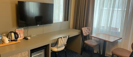 A hotel room with a flat-screen TV, a small desk with a kettle, a chair, a table, and a mini-fridge.
