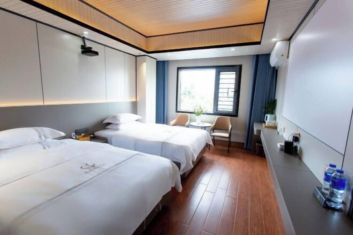 A hotel room with two beds, a small table, a chair, a window with curtains, and a water dispenser.