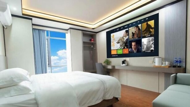 A hotel room with a bed, a desk, a TV with movie posters, a window with curtains, and a view of the sky. - Mancheng Boutique Hotel (Mengla)