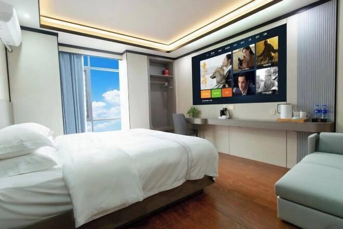 A hotel room with a bed, a desk, a TV with movie posters, a window with curtains, and a view of the sky.