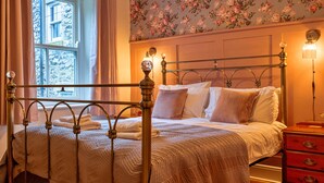 Un letto rifatto con ordine, con struttura in metallo, un comodino in legno con cassetti e una carta da parati con motivo floreale.