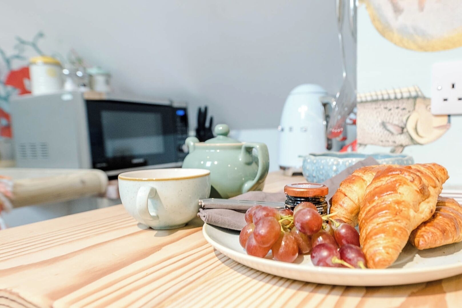Een ontbijtje met een bord croissants, druiven en jam, plus twee kopjes en een theepot op een houten tafel.
