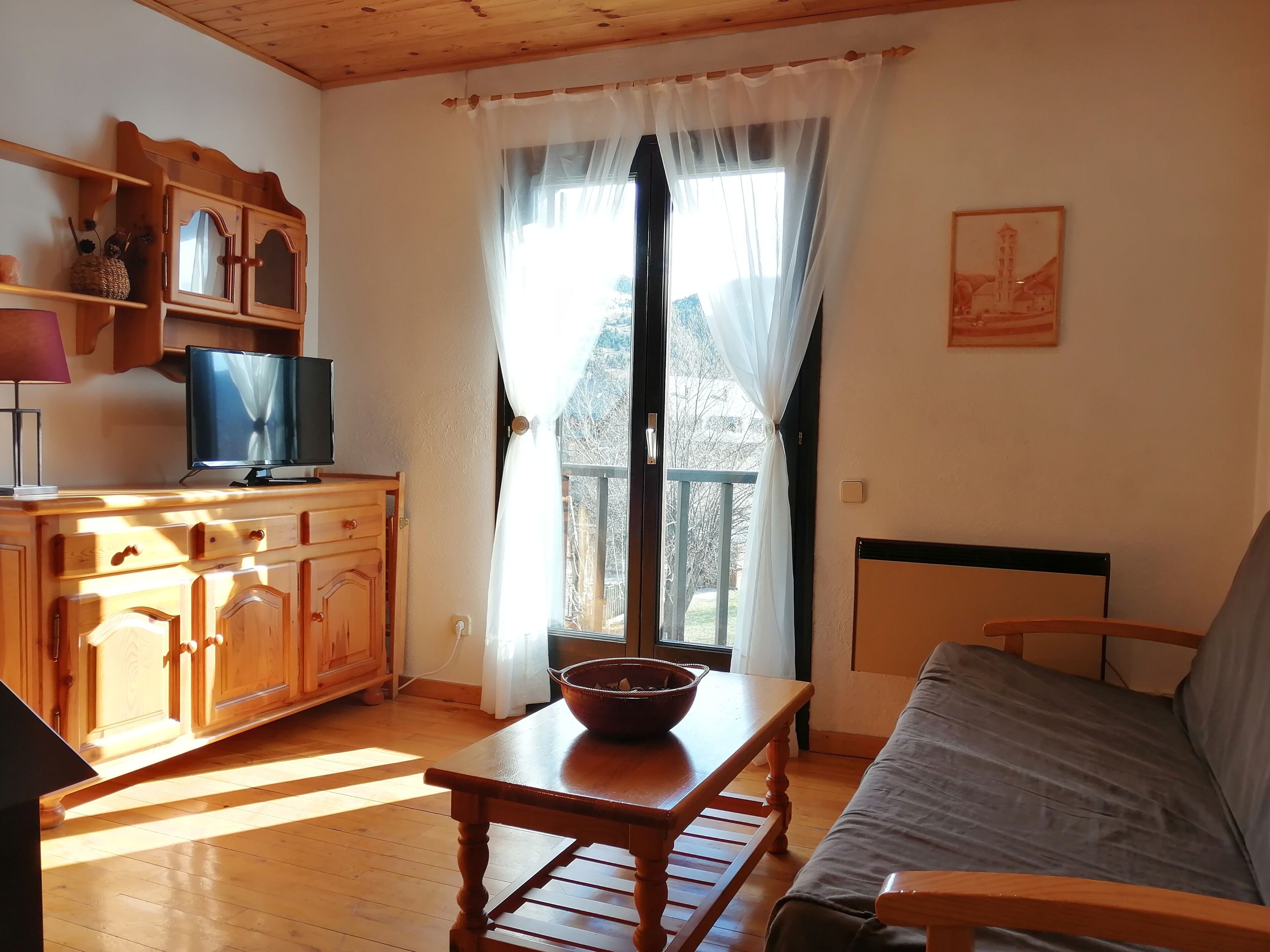 Une pièce comprenant un meuble TV en bois, un canapé, une table avec un saladier, une fenêtre avec des rideaux et un tableau accroché au mur.