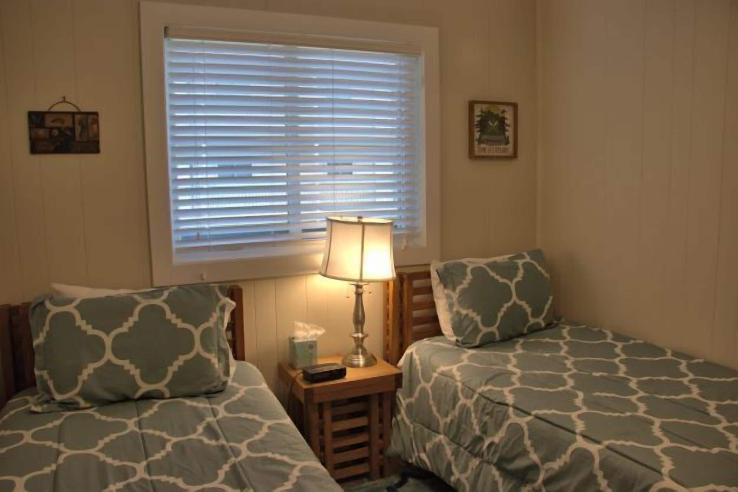 A bedroom with two beds, a nightstand with a lamp, and a window with blinds.