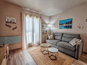 Un salon moderne avec un canapé gris, deux tables rondes, une grande fenêtre avec des rideaux légers et un tableau représentant un paysage enneigé.