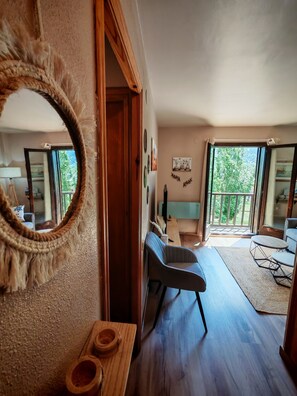 A spacious room with a wooden floor, a round mirror, a wooden bench, a chair, and a small table. There is a door leading to another room, and a window with a view of greenery.