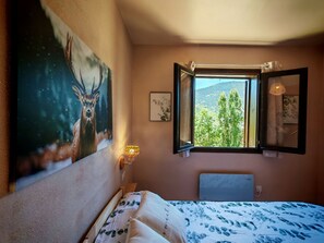 A bedroom with a bed, a painting of a deer, a window with an open shutter, and a wall-mounted lamp.