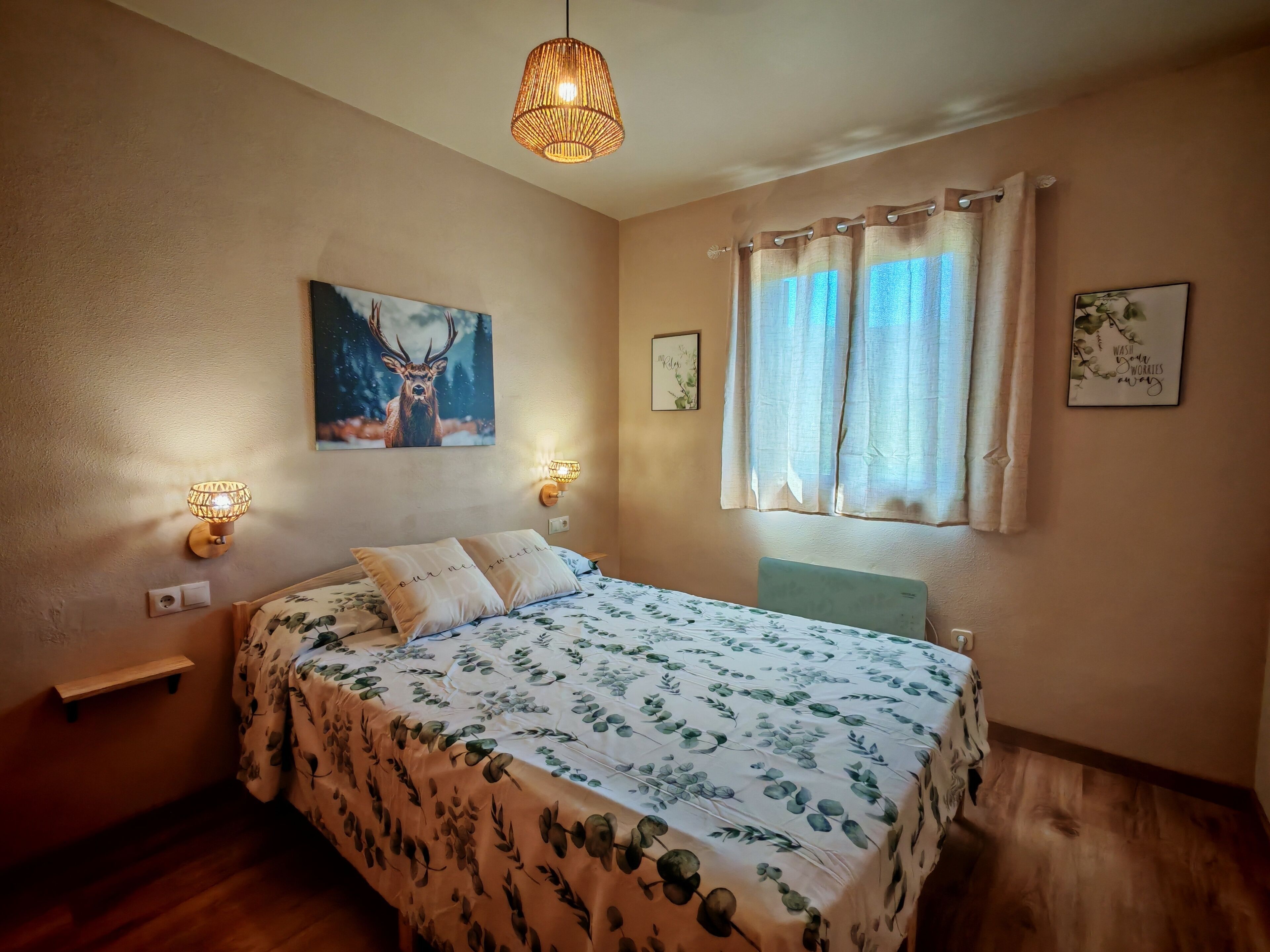 Een slaapkamer met een bed, twee wandlampen, een schilderij van een hert, een raam met gordijnen en een klein plankje.