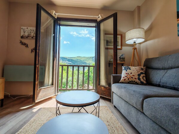 A living room with a sofa, a small table, a lamp, and a balcony with a view of hills.