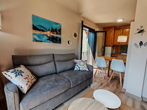Un salon moderne avec un canapé gris, des coussins à motifs, un tableau représentant un paysage enneigé, un coin repas avec une table et des chaises en bois, et une cuisine avec des meubles en bois.