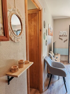 A room with a wooden door, a mirror with a fringe, a shelf with wooden containers, a grey upholstered chair, and a wall with framed artwork.