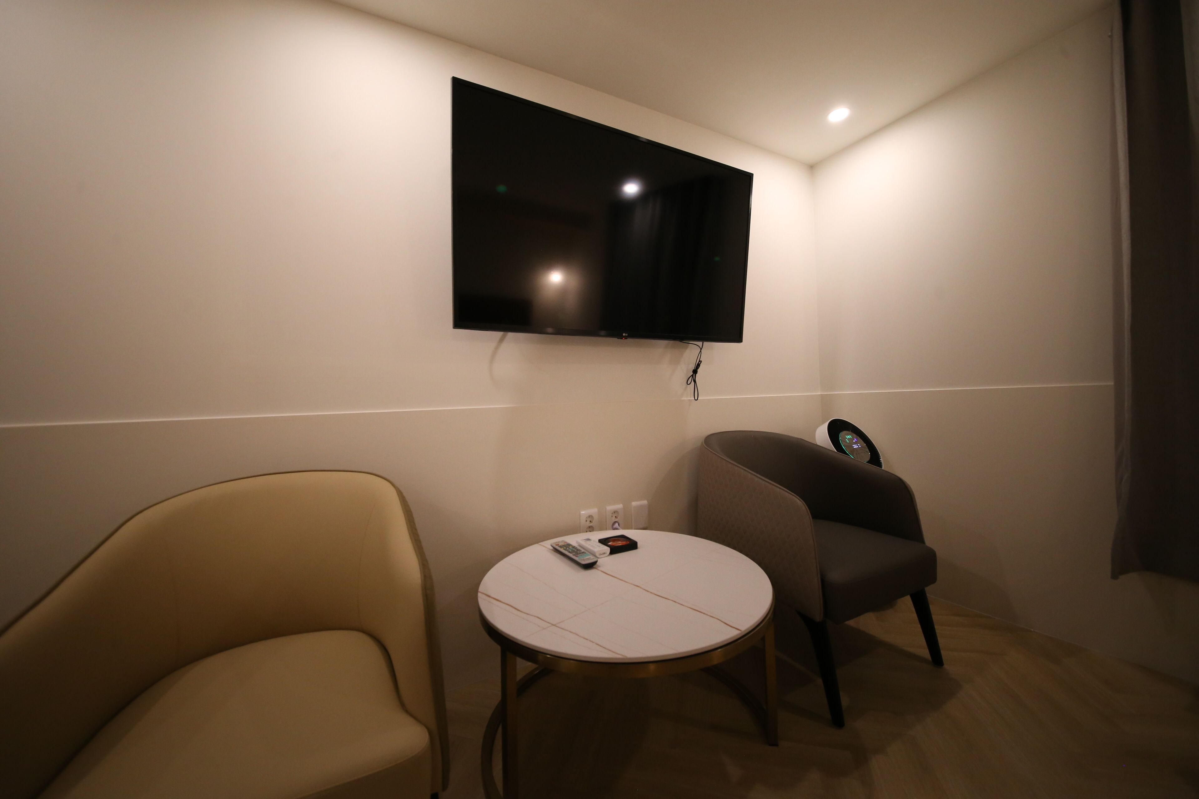 A room with a flat-screen TV mounted on the wall, a round table with a remote control, and two chairs.