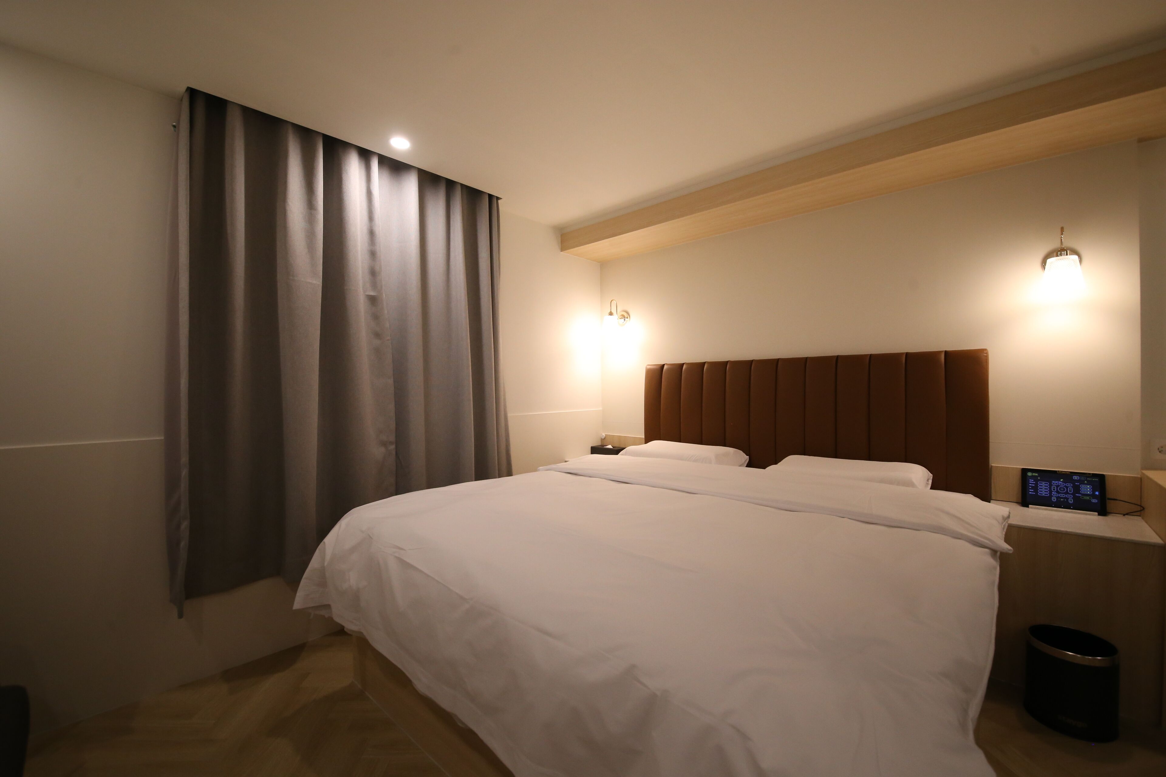 A hotel room with a large bed, wooden headboard, two bedside tables with lamps, a wall-mounted TV, and a window with curtains.