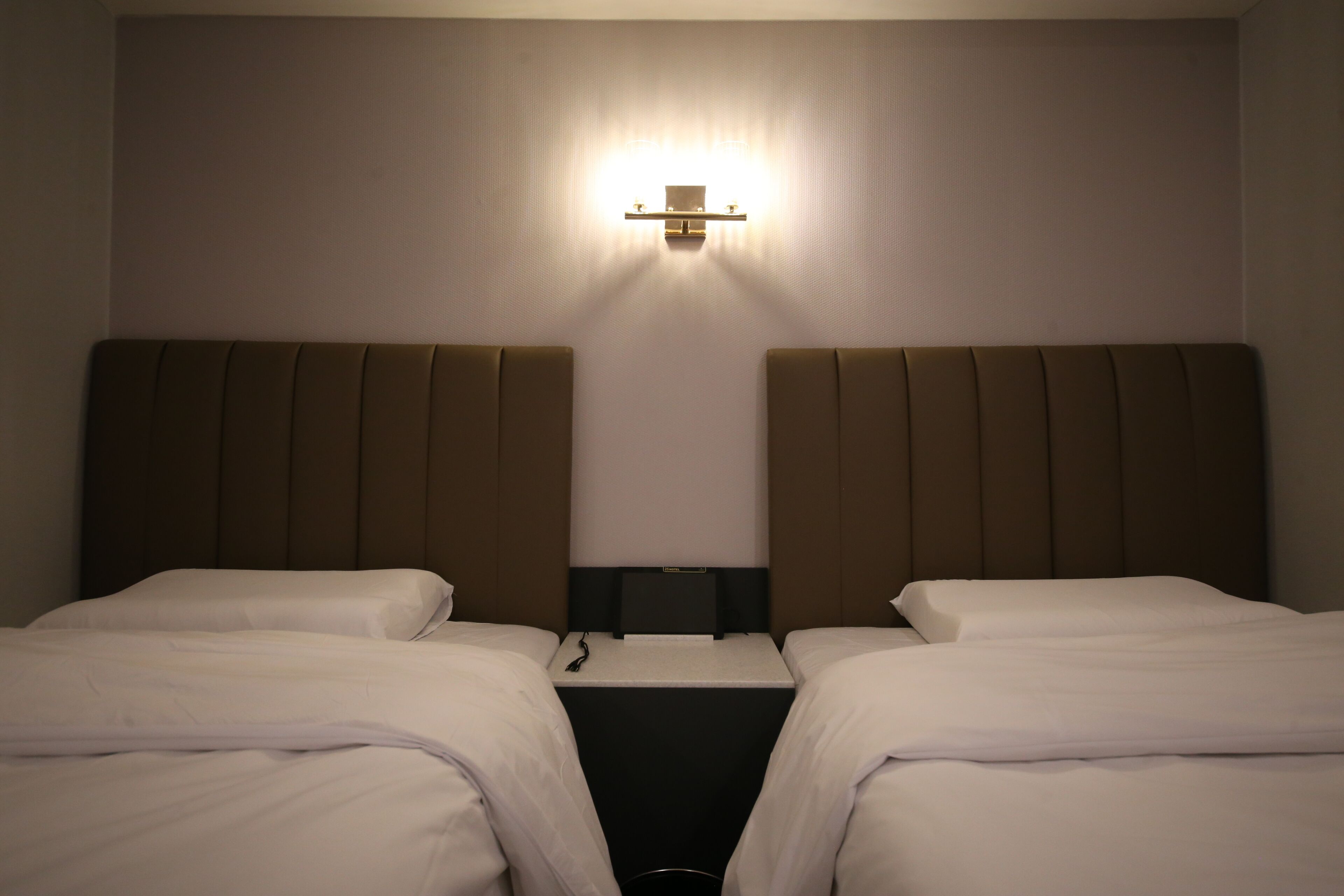 A hotel room with two beds, a nightstand, and a wall-mounted light fixture.