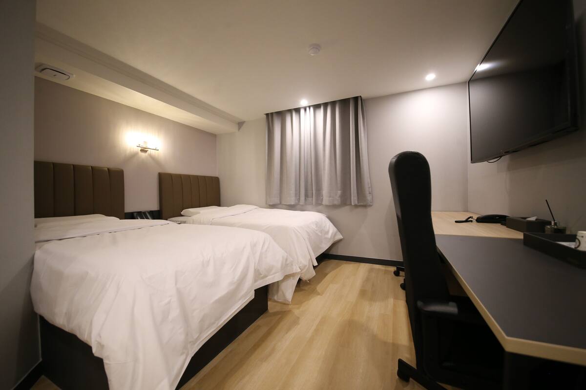Habitación de hotel con dos camas, un escritorio, una silla, un televisor de pantalla plana y una ventana con cortinas.