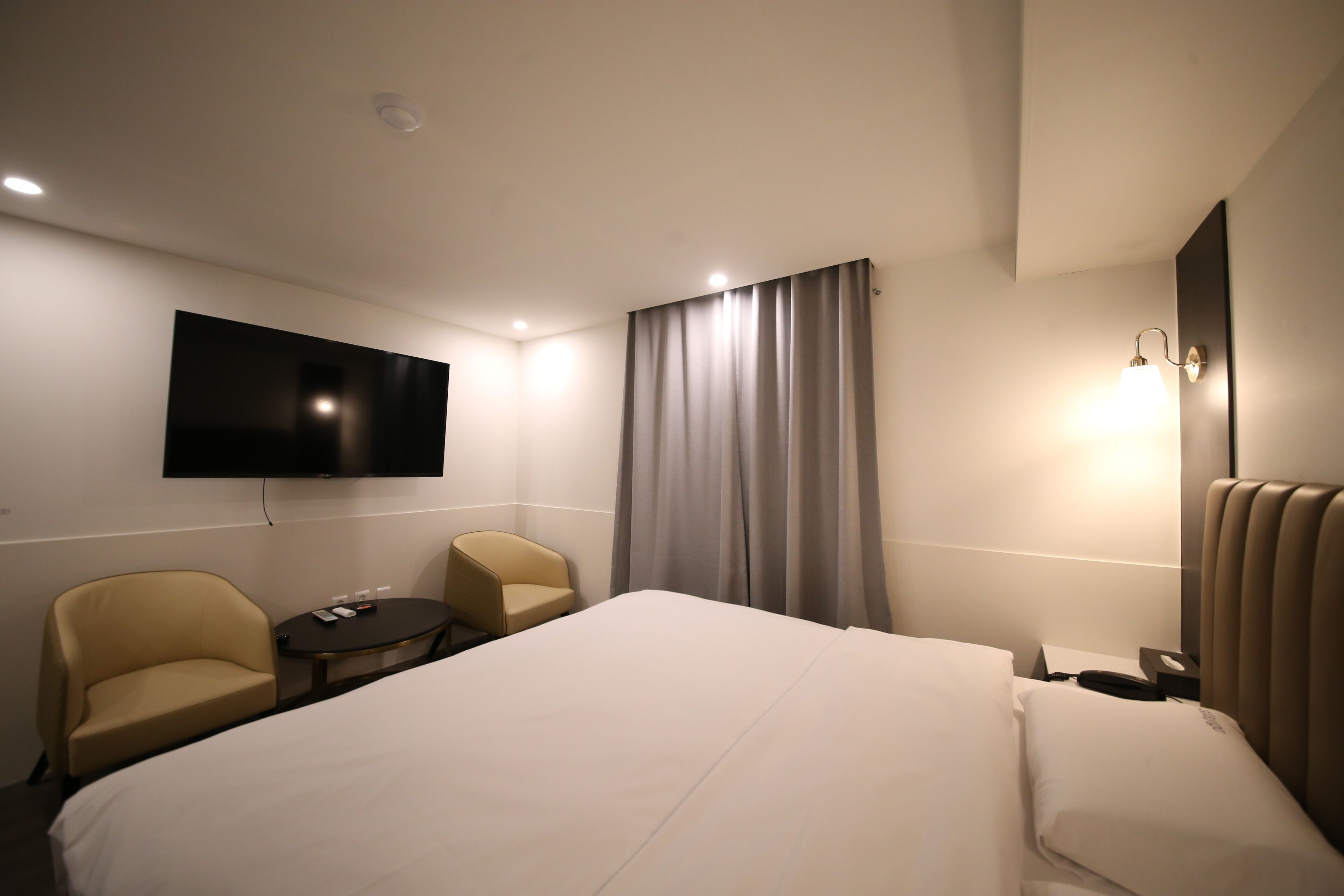 A hotel room with a large bed, two armchairs, a small table, a flat-screen TV, a bedside lamp, and a window with curtains.